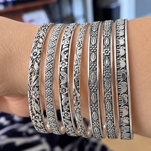Puede incluir: Un conjunto de siete brazaletes de metal plateado con varios patrones en relieve, incluyendo motivos florales, formas geométricas repetitivas, peces estilizados y una fila de figuras de elefantes, con un acabado oxidado.