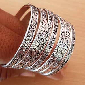 Puede incluir: Un conjunto de siete brazaletes de metal plateado con intrincados patrones en relieve que incluyen flores, pájaros, líneas onduladas, lunas crecientes y estrellas. Cada pulsera tiene un diseño texturizado distinto con detalles oxidados.