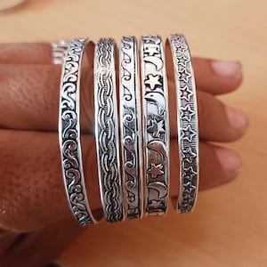 Pode incluir: Um conjunto de cinco pulseiras de metal prateado com padrões em relevo distintos. Os designs incluem volutas, uma textura de corda trançada, linhas onduladas repetitivas e uma série de luas crescentes e estrelas.