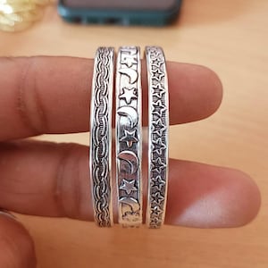 以下が含まれることがあります： 異なるエンボス加工が施された3つのシルバー調の金属製リング。左のリングは編み込み模様、中央のリングは三日月と星の連続模様、右のリングは小さな五芒星が繰り返されるデザインが特徴です。