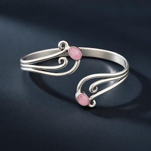 Könnte beinhalten: Ein silberner Armreif mit offenem Design, bestehend aus geschwungenen Metalllinien, die in zwei glatten, ovalen rosa Steinen enden. Der Armreif hat eine polierte metallische Oberfläche und eine geschwungene, flexible Struktur mit Öffnung.