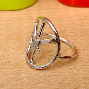 May include: A silver ring with a star design. The ring features a teardrop-shaped frame with a star in the center. The ring is set on a wooden surface, with a blurred green and red background.