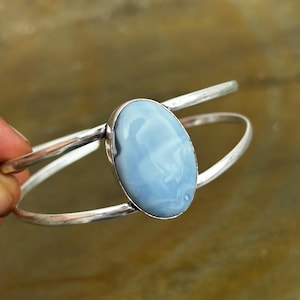 Peut inclure: Un bracelet manchette en argent orné d'une pierre ovale bleu clair au centre, dotée d'une surface lisse et polie. Le bracelet est composé de deux fils d'argent fins et parallèles qui se rejoignent au niveau du sertissage de la pierre.