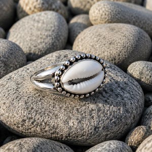 May include: A silver ring featuring a central white cowrie shell set within a beaded metal border. The band of the ring is split into two distinct silver segments that meet at the sides, creating a polished and metallic finish for the jewellery piece.