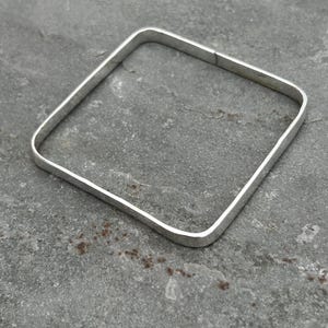 May include: A square-shaped bangle bracelet with a smooth, polished silver-toned finish. The bangle features rounded corners and a continuous, solid band construction, presenting a clean and uniform appearance throughout the entire piece of jewelry.