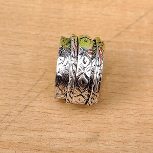 May include: A silver spinner ring with detailed patterns. The ring has multiple bands, each with a unique design, including floral and geometric motifs. The ring is displayed on a light brown wooden surface.