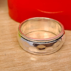 May include: A silver ring with a hammered texture and a spinning band. The ring sits on a wooden surface, with a red object in the background. The ring's design is visible.
