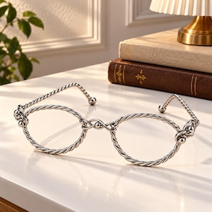 May include: A pair of silver-toned metal glasses featuring a twisted rope-like texture on the frames and temples. The design includes circular rims connected by a central bridge, with small spherical accents at the hinges and the ends of the temples.
