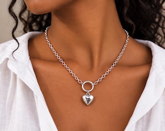 May include: A silver-toned necklace featuring a thick chain with round links and a central circular ring connector. A smooth, polished, puffed heart-shaped pendant hangs from the ring, resting against a white collared shirt.