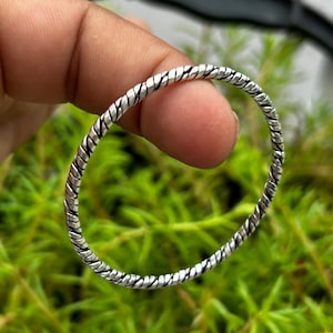 Puede incluir: Una pulsera tipo brazalete circular y delgada hecha de metal plateado. La superficie presenta una textura continua de cuerda trenzada, con surcos oscuros y profundos que crean un contraste marcado con los bordes pulidos y elevados.