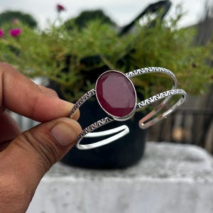 Puede incluir: Un brazalete de plata con una gran piedra preciosa de color rojo oscuro y forma ovalada en el centro. La banda de metal tiene un patrón geométrico repetitivo y texturizado en su superficie exterior y un diseño abierto para el ajuste.
