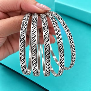 May include: A set of four silver-toned bangle bracelets, each featuring a continuous, repeating infinity-style knot pattern engraved along the outer surface. The bracelets have a polished finish with darkened recessed areas to highlight the design.