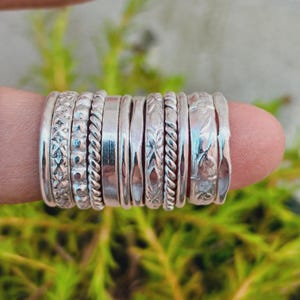 May include: A set of ten silver rings featuring various textures and patterns, including twisted rope designs, smooth bands, hammered finishes, and intricate engraved details, all arranged side by side in a stacked formation on a finger.