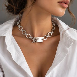 Puede incluir: Un collar de cadena en tono plateado que presenta una serie de eslabones circulares interconectados y un cierre de barra rectangular posicionado en la parte frontal. El collar descansa sobre el cuello, mostrando un acabado metálico pulido.