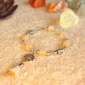 Puede incluir: Una pulsera con cuentas de color amarillo pálido ensartadas en alambre plateado. Un dije plateado con diseño de estrella y una pequeña borla colgando. La pulsera se exhibe sobre una superficie clara y texturizada.