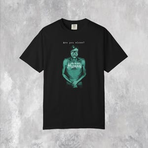 以下が含まれることがあります： 黒いTシャツ。水色の人物のグラフィックと、その上に「Are you alone?」というテキスト。グラフィックには「NO I'M NOT HUMAN」というテキストが含まれています。
