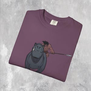 May include: Plum-colored t-shirt featuring a cartoon illustration of a muscular caveman with a spear chasing a dark gray dinosaur. The shirt has a crew neck and a tag in the upper right corner.