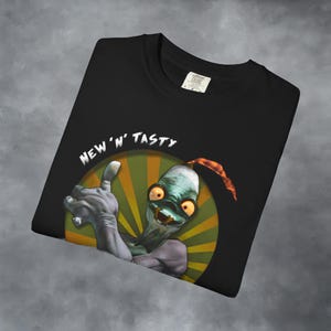 May include: Black t-shirt featuring a cartoon character with a green face, orange eyes, and a red antenna. The character is giving a thumbs up, with the words "NEW 'N' TASTY" above it. The design is set against a green and yellow circular background.