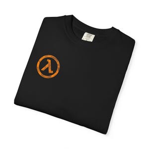 Könnte beinhalten: Schwarzes T-Shirt mit einem orangefarbenen Lambda-Symbol in einem Kreis auf der Vorderseite. Das Logo hat einen Used-Look. Das T-Shirt ist gefaltet und aus Baumwolle. Das Etikett zeigt "Comfort Colors".