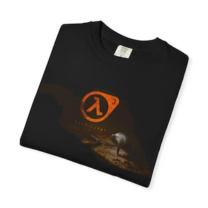 Könnte beinhalten: Schwarzes T-Shirt mit dem orangefarbenen Half-Life-Logo und der Zahl 3. Das Design beinhaltet eine Figur und die Worte "HALF-LIFE". Das T-Shirt hat einen Rundhalsausschnitt und ein Etikett am Kragen.