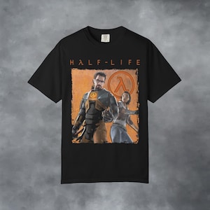 May include: A black short-sleeved t-shirt featuring a graphic print of the Half-Life video game characters Gordon Freeman and Alyx Vance. The design includes the orange Half-Life logo and the text HALF-LIFE printed in orange above the characters.