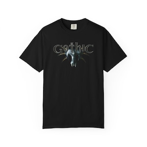 以下が含まれることがあります： 黒いTシャツ。銀色のスタイリッシュなフォントで「Gothic」の文字がプリントされ、その下にスカルのグラフィックがあります。スカルは暗くメタリックな外観で、複雑なディテールが施されています。Tシャツはクラシックなクルーネックです。