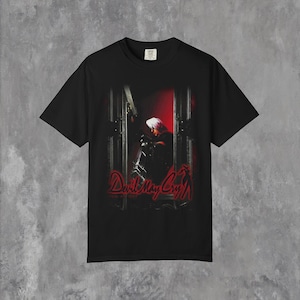 Könnte beinhalten: Schwarzes T-Shirt mit einer Grafik aus dem Videospiel "Devil May Cry". Das Design zeigt eine Figur mit weißen Haaren und das Logo des Spiels in Rot. Das T-Shirt hat einen Rundhalsausschnitt und kurze Ärmel.