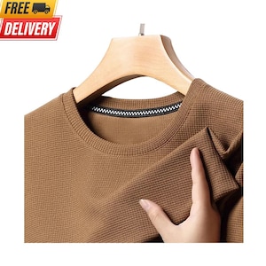 May include: A brown long-sleeve shirt with a textured waffle-knit fabric pattern. The garment features a round neckline with a black and white checkered trim detail along the inner collar, displayed on a light-colored wooden clothes hanger.