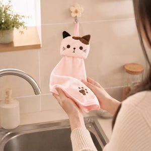 Könnte beinhalten: Ein rosa handtuchförmiges Handtuch mit einem braunen und schwarzen Katzengesicht-Design. Das Handtuch hat einen Pfotenabdruck auf der Unterseite und hängt an einem blütenförmigen Haken. Das Handtuch besteht aus einem weichen, plüschigen Material.
