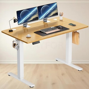 Puede incluir: Un escritorio de pie ajustable de color madera clara con dos monitores de ordenador, un teclado y un ratón. El escritorio tiene patas blancas y un panel de control. También se ve una alfombrilla negra, una lámpara pequeña y unos auriculares.