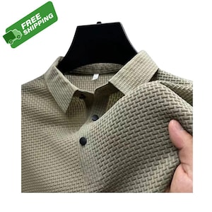 Puede incluir: Camisa verde oliva con textura tejida y botones negros. La camisa tiene un cuello clásico. Se ve una pancarta verde con "FREE SHIPPING". La tela de la camisa está siendo sostenida.