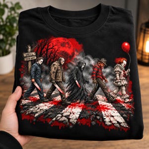 Puede incluir: Una camiseta negra con un estampado gráfico de cinco personajes de películas de terror caminando por un paso de cebra. La escena incluye una luna roja, humo y un letrero que dice HELL THIS WAY. Los personajes aparecen con cuchillos y un globo rojo.