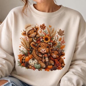 Op de afbeelding: Een crèmekleurig sweatshirt met ronde hals en een centrale afbeelding van drie Schotse hooglanders, omringd door herfstelementen zoals pompoenen, zonnebloemen, korenaren, dennenappels en herfstbladeren in oranje, bruine en groene tinten.