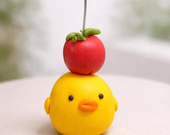 May include: A whimsical, handmade figurine featuring a bright yellow chick with a red apple on its head. The chick has small black eyes, an orange beak, and tiny ear-like protrusions. A silver wire extends from the apple, adding a decorative touch.