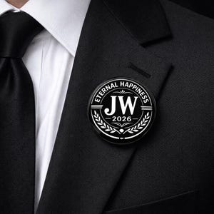 May include: A circular black pin featuring white text that reads ETERNAL HAPPINESS, the initials JW, and the year 2026. The design includes a decorative laurel wreath motif and horizontal lines, pinned to the lapel of a black suit jacket.