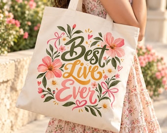 May include: A cream-colored canvas tote bag featuring the text Best Live Ever in dark green, yellow, and pink script lettering. The design is surrounded by a floral arrangement of pink and white flowers, green leaves, and small pink hearts.