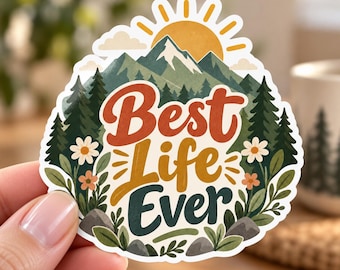 May include: A die-cut sticker featuring a mountain landscape with a yellow sun, green pine trees, and white flowers. The text "Best Life Ever" is printed in the center in orange, yellow, and dark green script and block lettering on a white background.