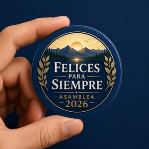 Puede incluir: Un pin circular azul con un paisaje de monta&ntilde;a al amanecer, un bosque y un lago. El texto FELICES PARA SIEMPRE, ASAMBLEA y 2026 aparece en letras doradas. Dos ramas de laurel doradas enmarcan el texto central sobre un fondo azul oscuro.