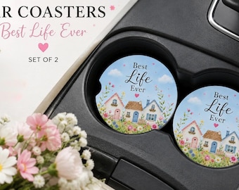 May include: Two round car coasters featuring a light blue sky background with three small houses, colorful flowers, and the text Best Life Ever. The coasters are placed in a black vehicle cup holder, accompanied by white and pink flowers nearby.