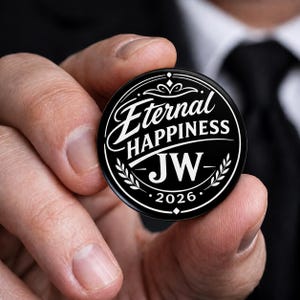 May include: A circular black pin-back button featuring white text that reads Eternal HAPPINESS JW 2026. The design includes a decorative border with stylized laurel leaves and geometric accents, creating a clean, high-contrast graphic appearance.