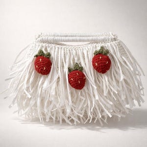 May include: A white, fringed handbag with three crocheted red strawberries with green leaves. The bag has a woven handle and a fringe made of white paper strips. The strawberries are evenly spaced across the front.