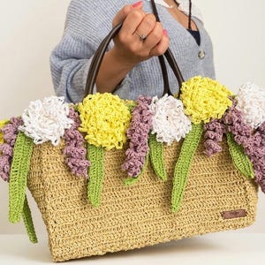 Könnte beinhalten: Beige gewebte Tragetasche mit braunen Ledergriffen, verziert mit gehäkelten Blumen in Weiß, Gelb und Lila. Grüne gehäkelte Stiele und Blätter ergänzen das Blumenarrangement. Ein kleines braunes Etikett ist an der Tasche befestigt.