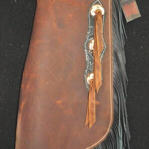 May include: Brown leather chaps with black fringe and silver embellishments. The chaps have a black leather top with embossed patterns and a belt with a silver buckle. A tag is attached.
