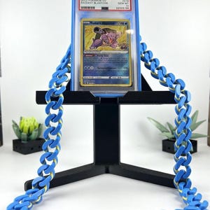Może przedstawiać: Karta Pok&eacute;mon Radiant Blastoise w niebieskim etui, wystawiona na czarnym stojaku. Karta zawiera ilustrację Blastoise. Niebieski łańcuch z ż&oacute;łtymi akcentami jest przerzucony przez stojak.