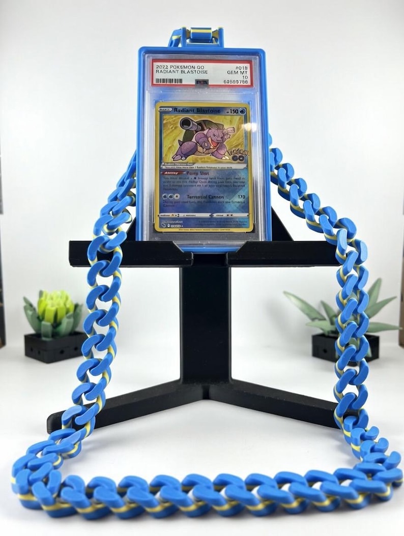 Może przedstawiać: Karta Pok&eacute;mon Radiant Blastoise w niebieskim etui, wystawiona na czarnym stojaku. Karta zawiera ilustrację Blastoise. Niebieski łańcuch z ż&oacute;łtymi akcentami jest przerzucony przez stojak.