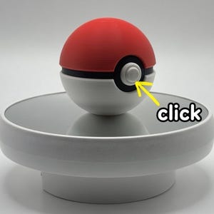 Könnte beinhalten: Ein rot-weißer Pokéball mit schwarzem Band und Knopf. Der Ball steht auf einem weißen, kreisförmigen Ständer. Das Wort "click" steht auf dem Ständer.