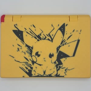 May include: Yellow handheld gaming console featuring a black Pikachu design. The console has a red accent on the top left corner and yellow hinges. The Pikachu graphic is stylized with a burst effect, giving it a dynamic appearance.