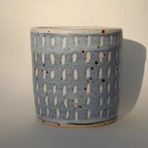May include: A ceramic tumbler with a light blue glaze and a pattern of white dashes. The cup has a textured surface and a white rim. Small brown speckles add detail. A unique piece for the home, suitable for holding approximately 300 ml.
