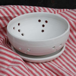 May include: A white ceramic colander bowl with drainage holes, sitting on a matching saucer. The bowl and saucer are on a red and white striped fabric.