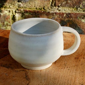 May include: A handcrafted, cream-coloured ceramic mug with a rounded form and a robust handle. The mug rests on a wooden surface, with a brick wall in the background. The mug's interior is shadowed, emphasising its depth and shape.